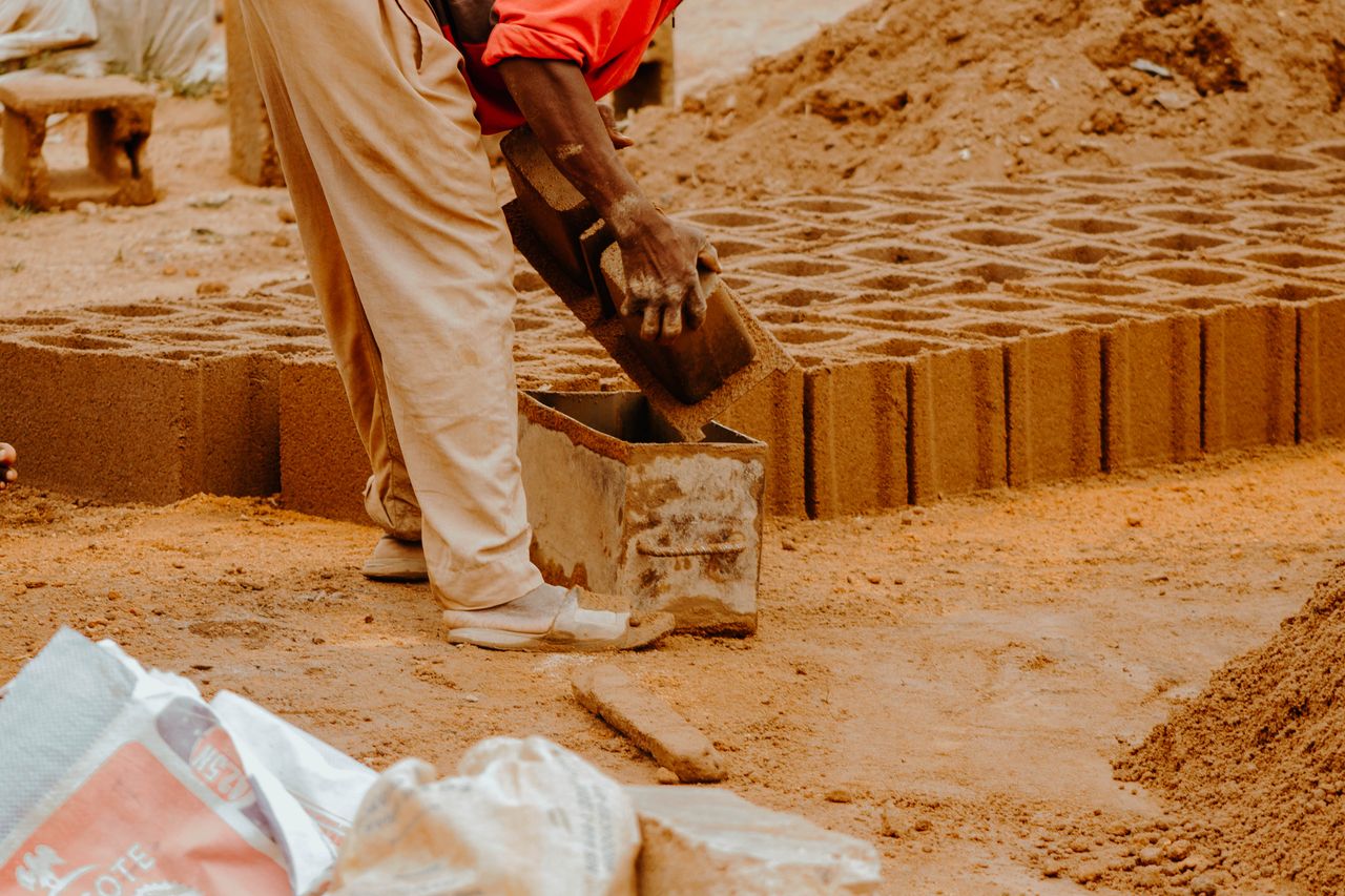 A worker shaping bricks with a concrete mold at an outdoor construction site.